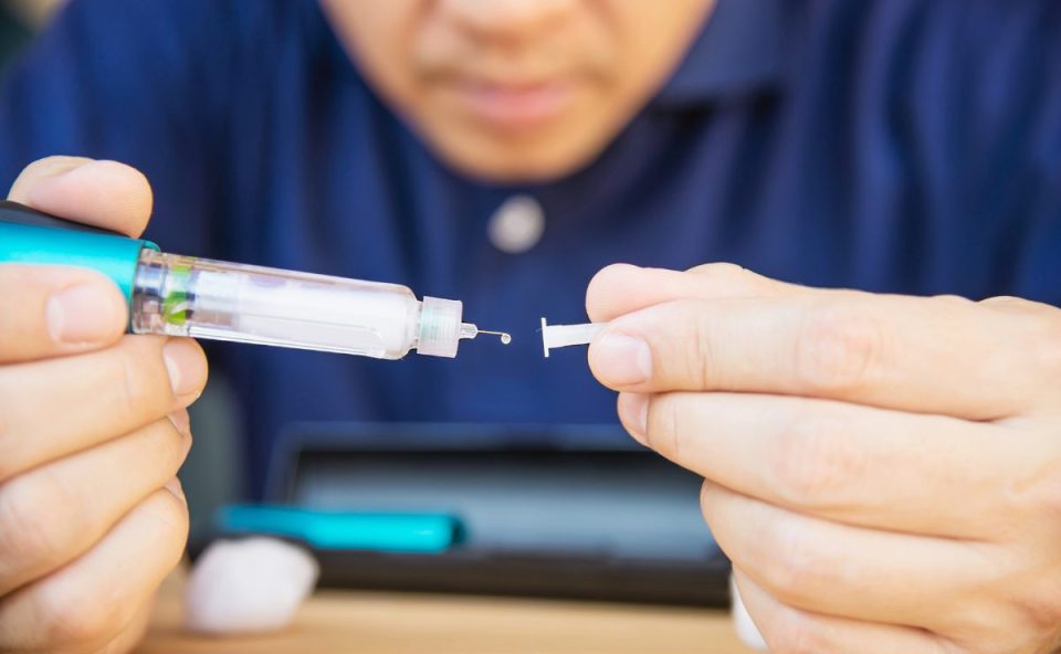 Homem em close-up segurando uma caneta de aplicação de medicamento injetável, removendo a tampa da agulha. Ao fundo, uma caixa de transporte e algodão.