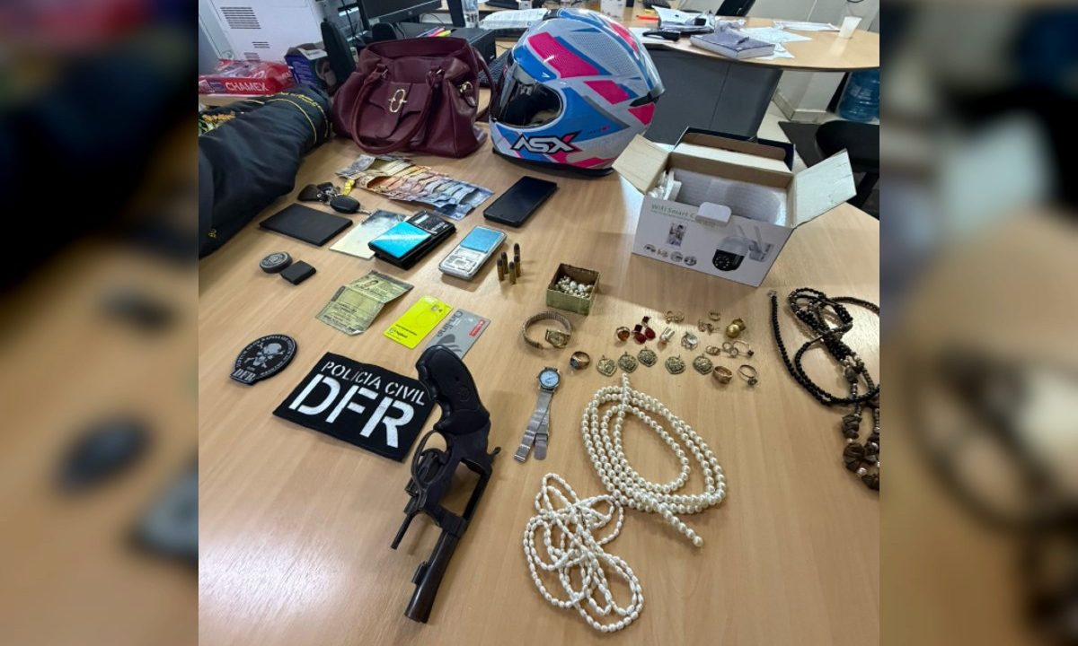 Joias, dinheiro, bolsa, celular, relógio, capacete de moto e outros objetos apreendidos, organizados sobre uma mesa em delegacia após a prisão de um suspeito de aplicar golpe contra uma idosa e levar seus pertences, incluindo joias.