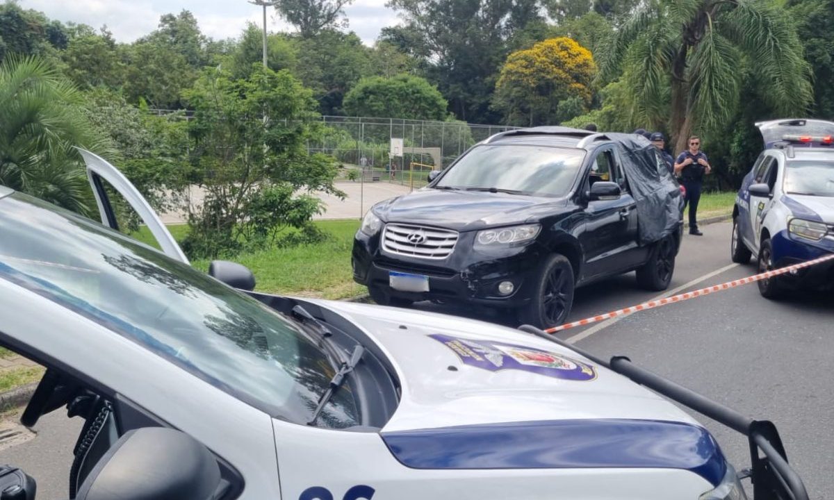 Imagem mostra uma viatura da Guarda Municipal ao lado do carro onde um homem foi encontrado morto no Parque Atuba. O veículo preto está com o vidro quebrado.