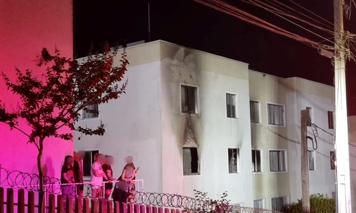 Foto mostra fachada externa do apartamento com marcas após incêndio no bairro Campo Comprido, em Curitiba. A parede, que é branca, agora tem sinais pretos por causa do fogo. Moradores aparecem do lado de fora