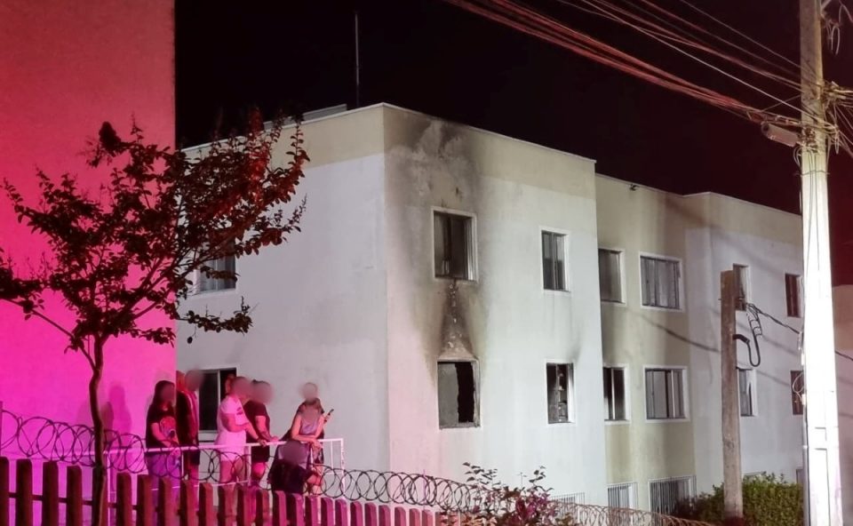 Foto mostra fachada externa do apartamento com marcas após incêndio no bairro Campo Comprido, em Curitiba. A parede, que é branca, agora tem sinais pretos por causa do fogo. Moradores aparecem do lado de fora