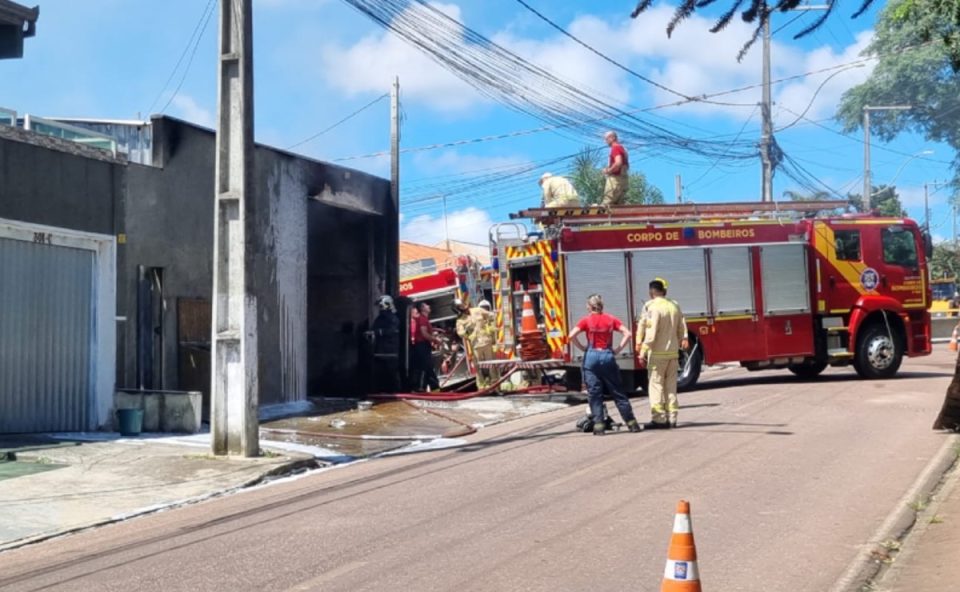 Bombeiros combatem incêndio em depósito de cotonetes em Piraquara, fachada do imóvel destruída e área isolada na rua.