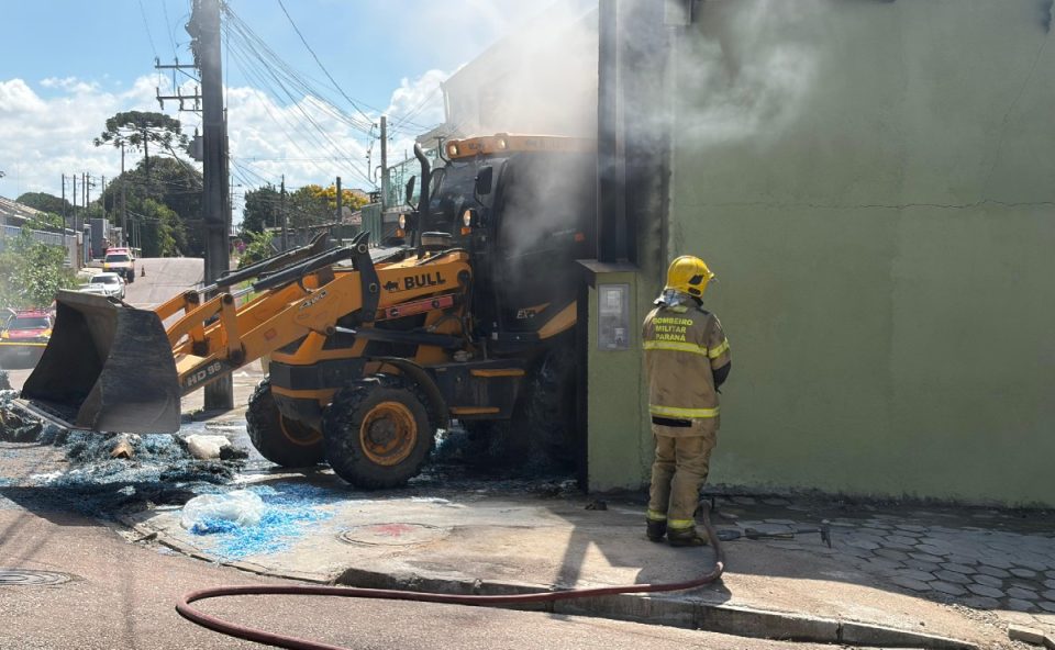 Máquina remove parte da carga de cotonete enquanto bombeiro realiza o rescaldo após incêndio em depósito em Piraquara, com fumaça saindo do imóvel.