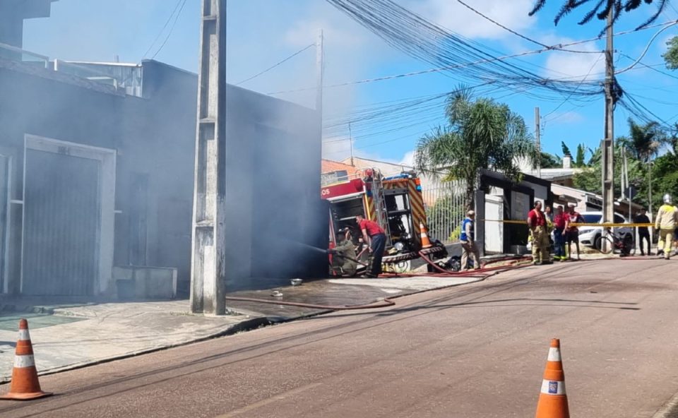 Bombeiros combatem incêndio em depósito de cotonetes em Piraquara, com fumaça saindo do imóvel e área isolada na rua.
