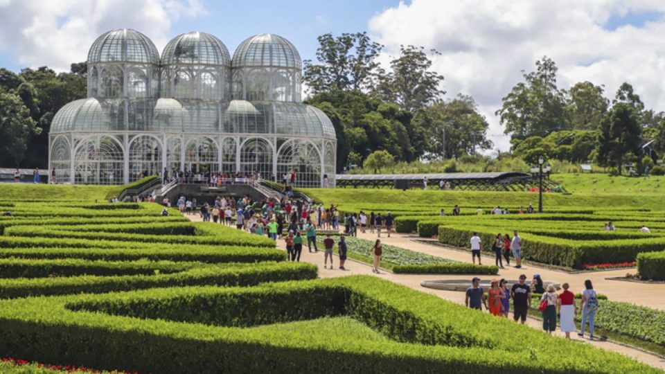 Vista do Jardim Botânico de Curitiba com estufa de vidro ao fundo e visitantes caminhando entre jardins geométricos bem cuidados.