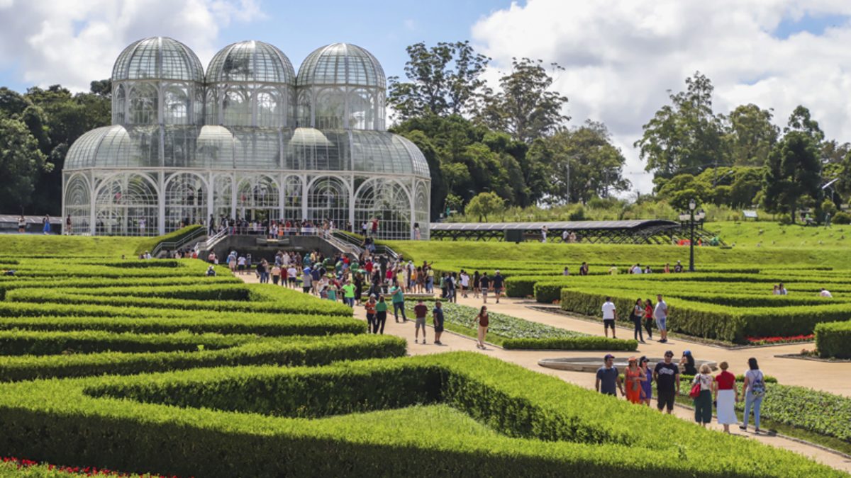 Vista do Jardim Botânico de Curitiba com estufa de vidro ao fundo e visitantes caminhando entre jardins geométricos bem cuidados.
