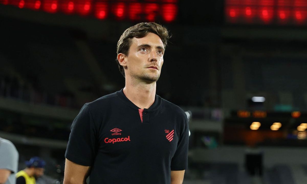 João Correia no Athletico aparece em pé à beira do campo, vestindo camisa preta do clube com detalhes em vermelho. O treinador olha para frente com expressão séria, em um estádio iluminado, com arquibancadas e luzes desfocadas ao fundo.