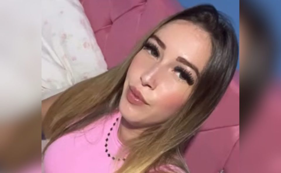 Imagem mostra a jovem assassinada dentro de um condomínio em São José dos Pinhais. Ela veste uma blusa rosa e usa um colar. Ela tinha cabelo castanho claro. Aparece sentada em uma cama com cabeceira cor de rosa também. Ela usa cílios postiço e faz bico para a foto. Está com cabelo solto.