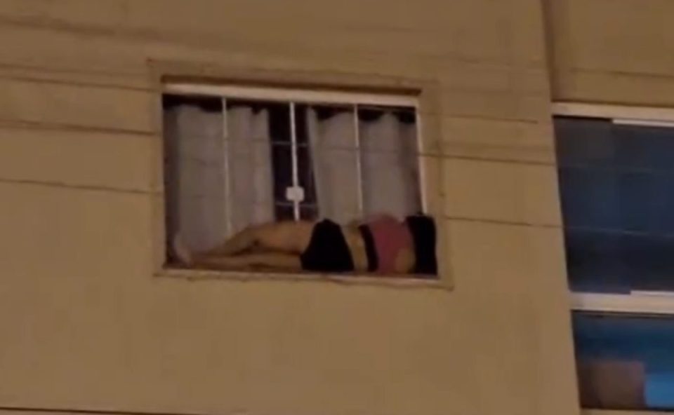 Imagem mostra uma jovem de shorts preto e blusa rosa deitada na janela de um apartamento no terceiro andar. O prédio é amarelo. A janela de vidro está fechada.