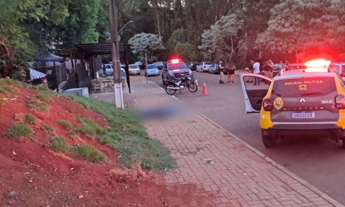 Foto tirada no início do amanhecer mostra um corpo coberto por saco plástico sobre uma calçada. O corpo está borrado por edição. Ao lado, há policiais militares fardados conversando com mulher. Perto deles há uma viatura da PM.