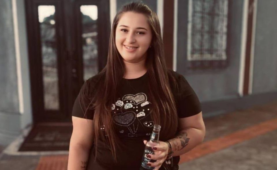 Foto mostra a jovem Jamily Cordeiro, de 21 anos, segurando uma garrafa de bebida em uma das mãos e sorrindo para foto. Ela tem cabelos castanho escuro, longos e usa aparelho nos dentes. A camiseta é preta e tem estampa de corações com brilhos. Jovem foi morta e teve a casa incendiada pelo suspeito
