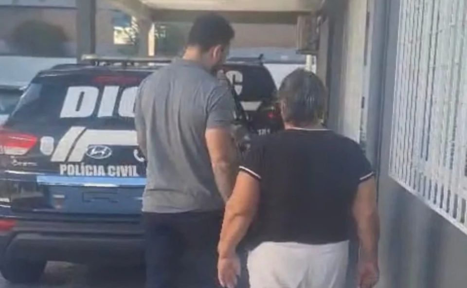 Foto mostra madrasta suspeita de matar enteada de costas e sendo presa, próximo a uma viatura da Polícia Civil. Homem conduz ela para dentro da delegacia