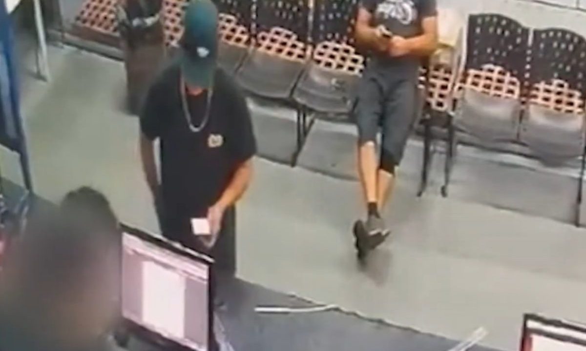 Imagem de câmera de segurança mostra dois homens dentro de uma loja; um deles, de boné e camiseta preta, está no balcão enquanto outro aguarda ao fundo, próximo a cadeiras.