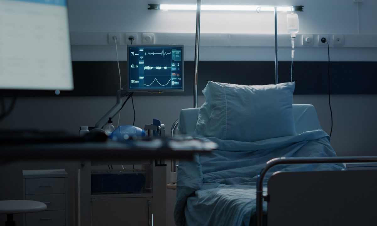 Leito hospitalar vazio em quarto com equipamentos médicos e monitor ligado, ilustrando a situação de retorno de funcionários em hospital de Curitiba após demissões sem pagamentos.