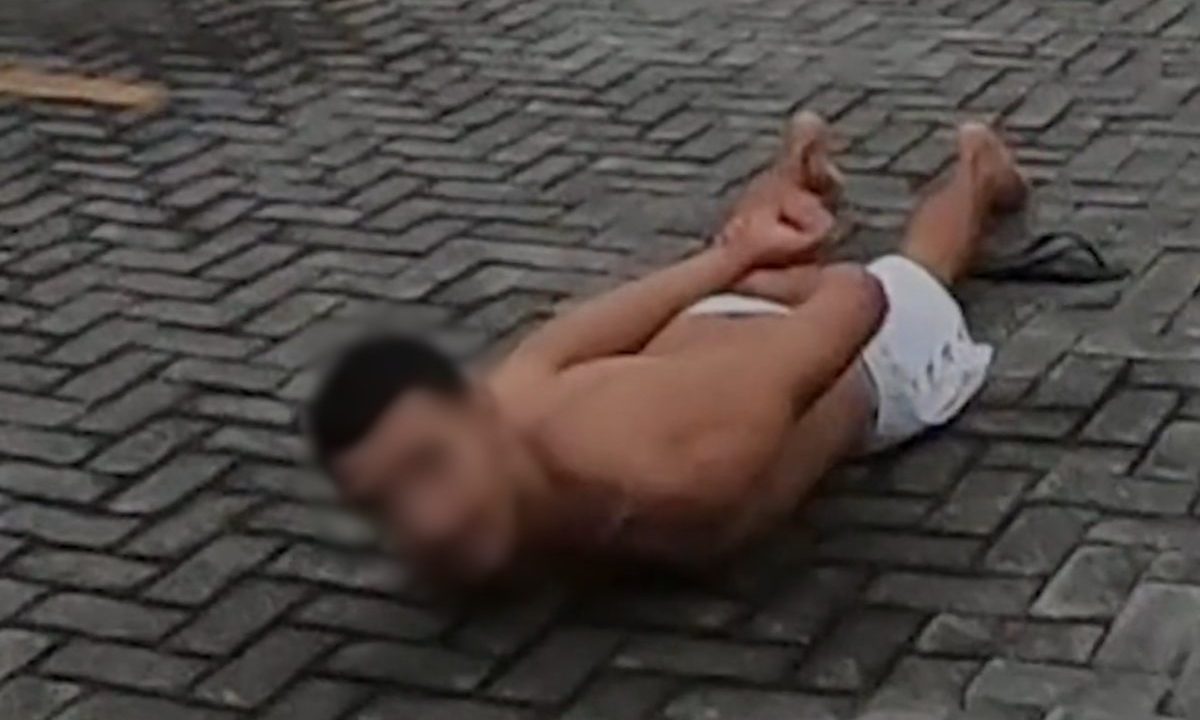 Mãe que chegou perto da morte ao ser brutalmente espancada pelo filho pede justiça. Imagem mostra o suspeito deitado de barriga para o chão. Ele usa bermuda branca. O rosto está borrado. O chão é de tijolos.