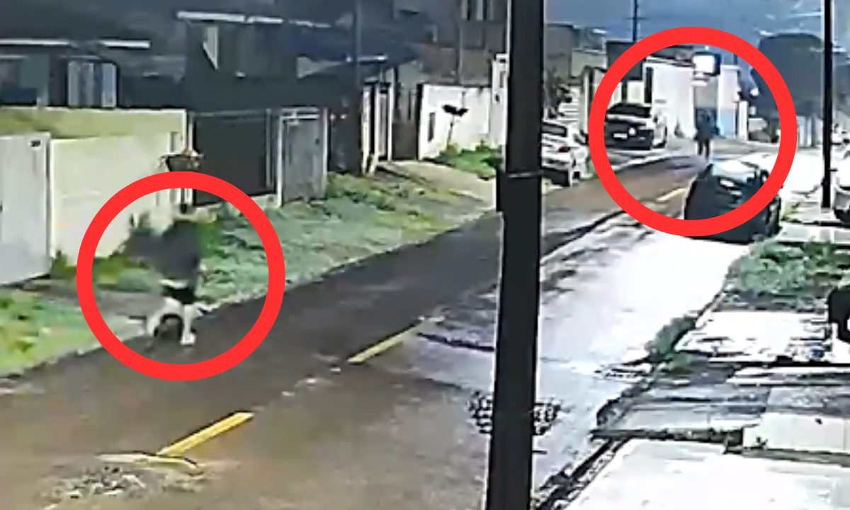 Mulher é perseguida por um homem em São José dos Pinhais. A imagem mostra o suspeito correndo em direção da vítima. A rua está vazia e molhada. Há carros estacionados perto das calçadas. O homem usa uma blusa de moletom e uma bermuda.