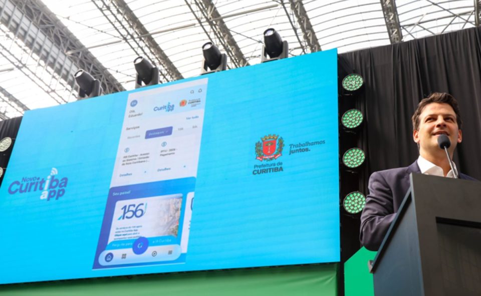 Prefeito de Curitiba apresenta o novo Curitiba App em telão durante evento, com interface do aplicativo exibida ao público.