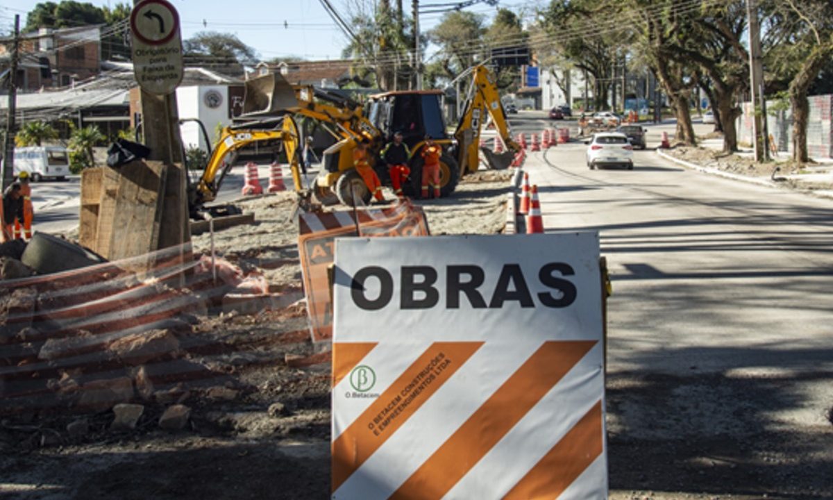 Placa de sinalização alerta para obras viárias com presença de máquinas em rua do bairro Mercês, em Curitiba, representando as intervenções do projeto Novo Inter 2.