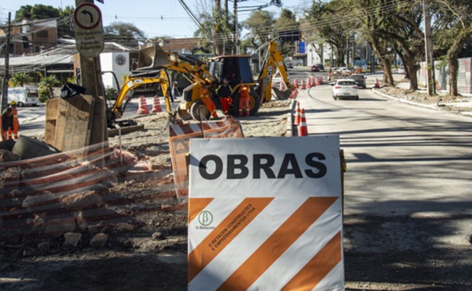 Placa de sinalização alerta para obras viárias com presença de máquinas em rua do bairro Mercês, em Curitiba, representando as intervenções do projeto Novo Inter 2.