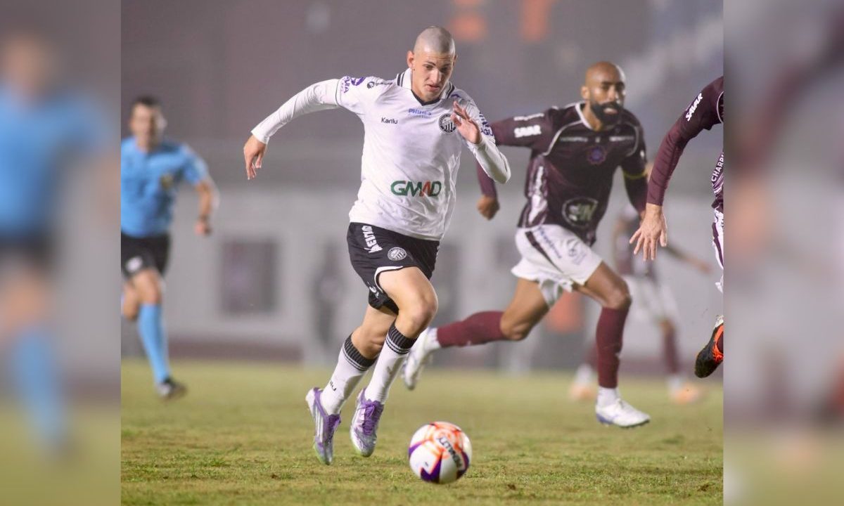 Em campo de futebol com gramado levemente desgastado e sob iluminação artificial, um jogador do Operário veste uniforme branco com detalhes pretos e corre em alta velocidade conduzindo a bola à frente dos pés. Ele tem cabelo raspado e está com expressão concentrada, inclinado para frente no movimento de arrancada. Logo atrás, dois adversários com uniformes em tom vinho tentam acompanhá-lo na disputa. Ao fundo, um árbitro com camisa azul observa a jogada. A cena transmite intensidade e disputa em um momento de transição ofensiva durante a partida.