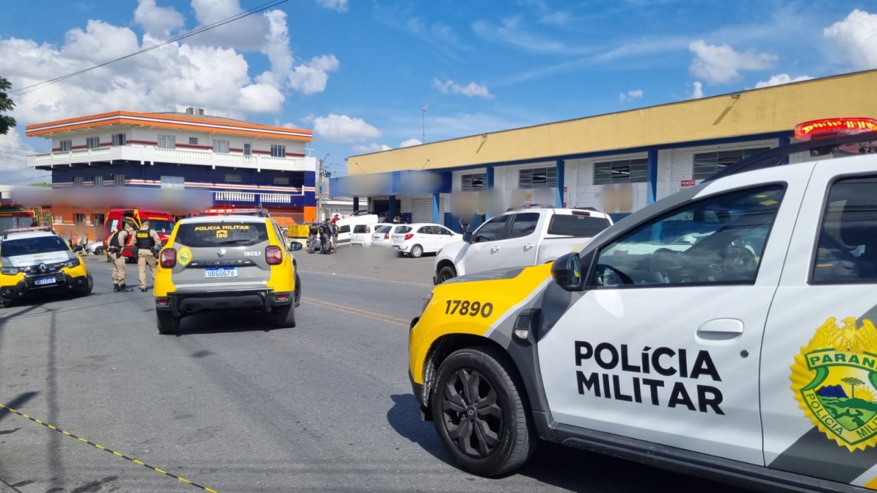 Viaturas da Polícia Militar em frente ao mercado onde pai e filho foram mortos a tiros em Almirante Tamandaré; ao lado de um carro, o corpo de uma das vítimas aparece caído e borrado na imagem.