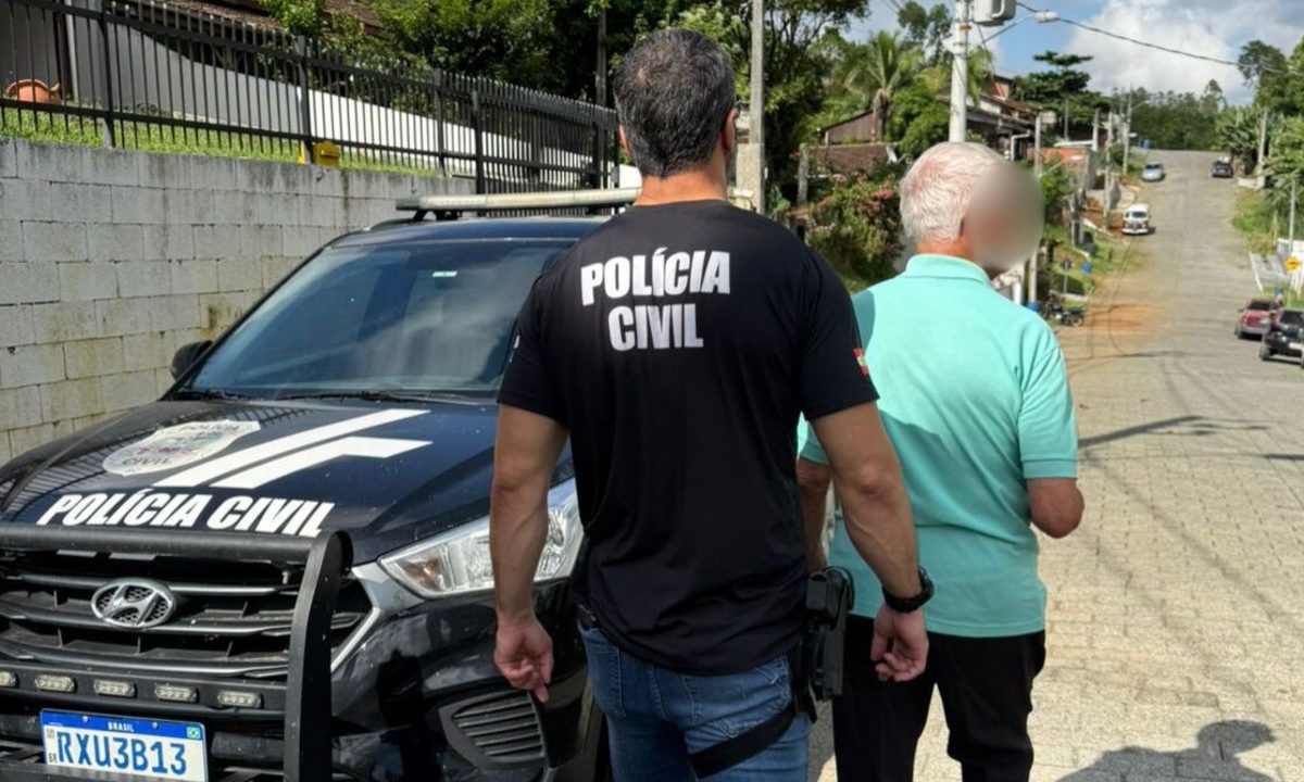Foto mostra pastor de 82 anos, que abusou de criança mais de 140 vezes durante seis anos, sendo preso por policial civil em SC. Condenado usa camiseta azul piscina e está de costas