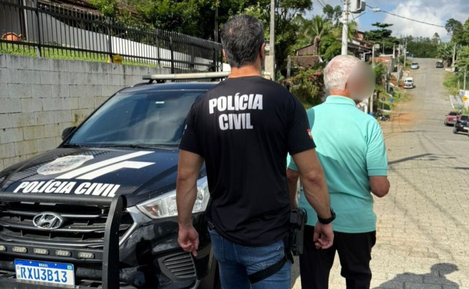 Foto mostra pastor de 82 anos, que abusou de criança mais de 140 vezes durante seis anos, sendo preso por policial civil em SC. Condenado usa camiseta azul piscina e está de costas