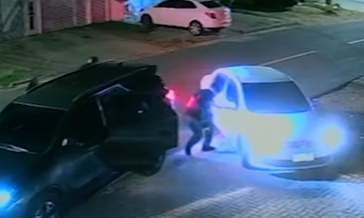Homem armado desce de veículo escuro e aborda motorista de carro branco durante a noite em rua residencial.