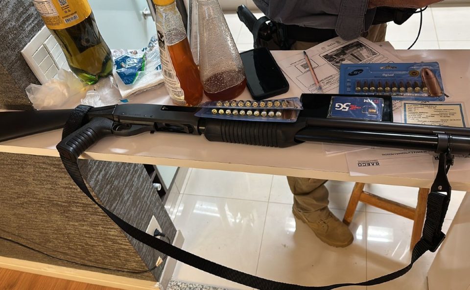 Arma de grosso calibre carregada e diversos itens apreendidos sobre uma mesa durante operação que teve policiais civis suspeitos de envolvimento com comércio ilegal de remédio para emagrecimento.