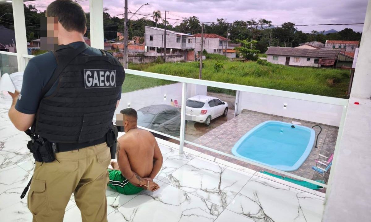 Foto mostra suspeito preso e policial do Gaeco em pé