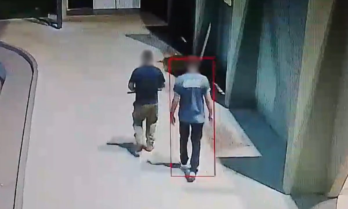 Prefeito afirma que homem em situação de rua agredido por chefe de vigilância ameaçava funcionários com faca. Imagem mostra o chefe de vigilância indo em direção à vítima. Ele segura um cacetete e usa calça bege e blusa azul marinho, está de costas. Ao lado, um servidor público efeitivo de calça jeans e camiseta azul.