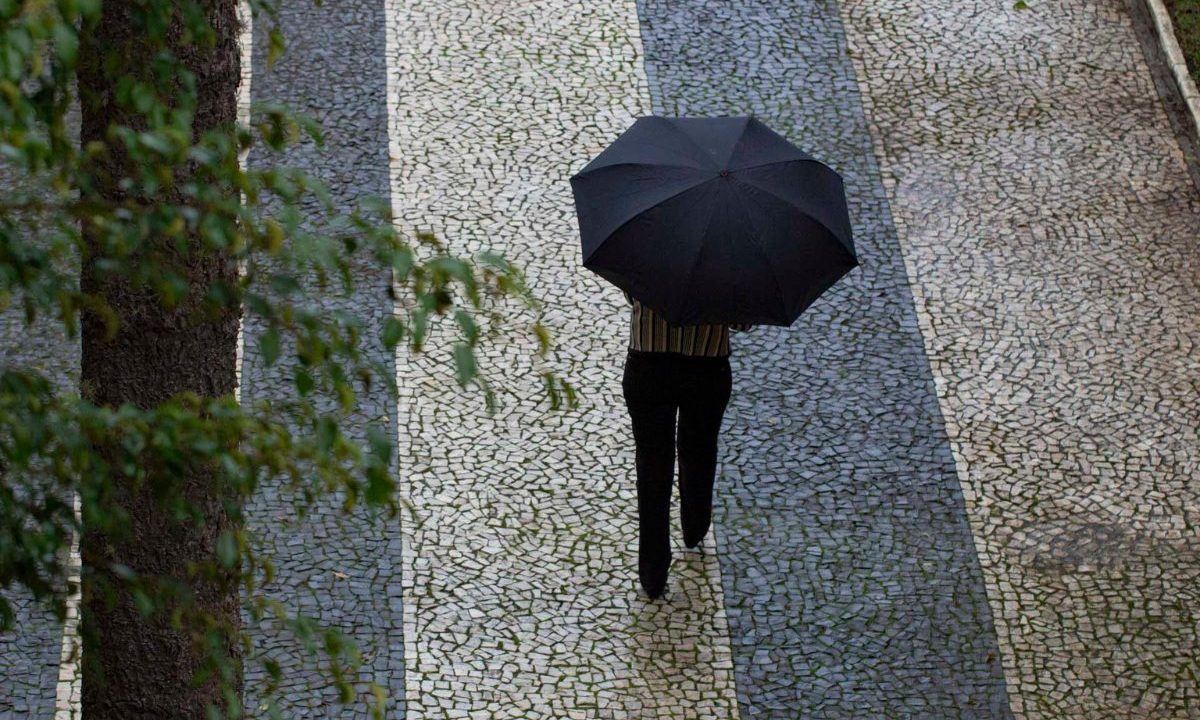 Imagem ilustrativa para a previsão do tempo em Curitiba nesta quarta-feira. Imagem mostra um dia chuvoso. Uma pessoa está caminhando em uma calçada segurando um guarda-chuva preto. Ela usa calça preta e blusa listrada.