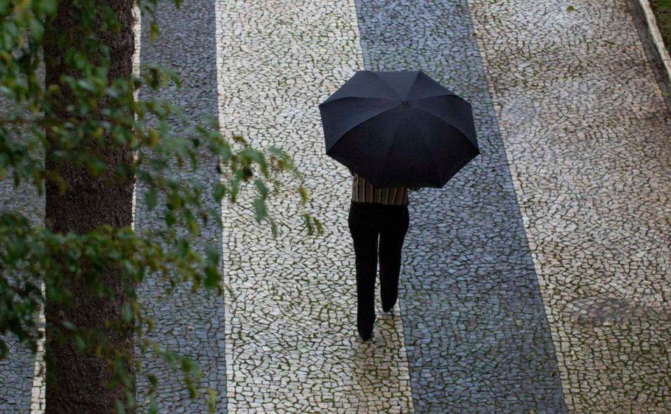 Imagem ilustrativa para a previsão do tempo em Curitiba nesta quarta-feira. Imagem mostra um dia chuvoso. Uma pessoa está caminhando em uma calçada segurando um guarda-chuva preto. Ela usa calça preta e blusa listrada.