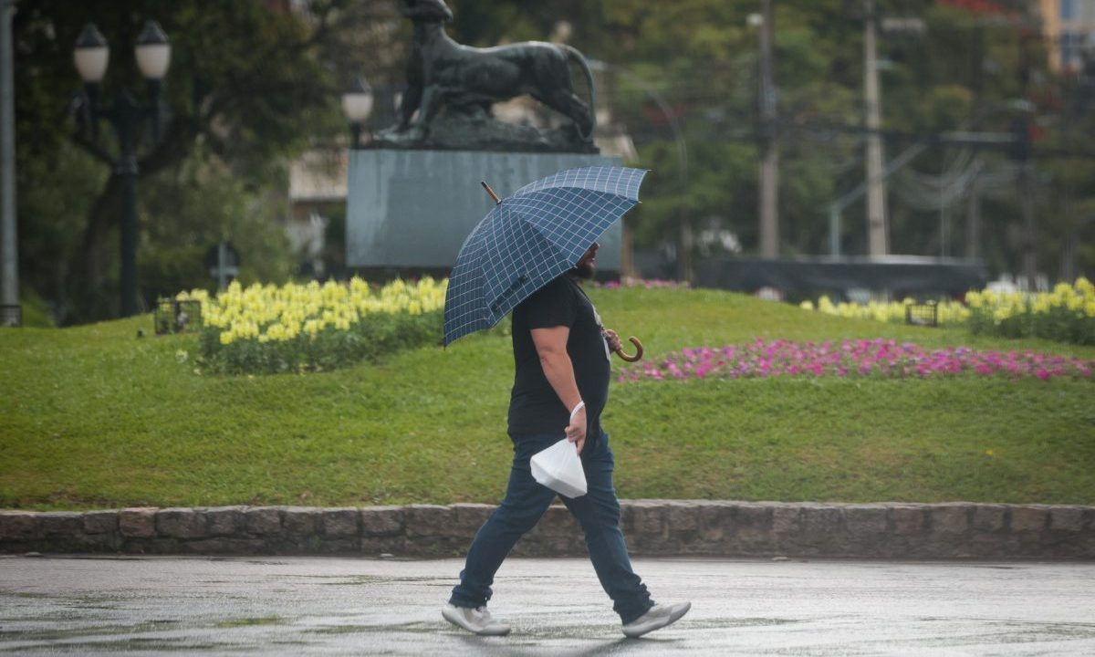 Imagem ilustra a previsão do tempo em Curitiba nesta terça-feira (30). Um homem de camiseta preta e calça jeans está caminhando na calçada, ao lado de um gramado verde, segurando um guarda-chuva aberto sobre a cabeça. O dia está chuvoso. Ele segura uma sacola branca na mão direita.