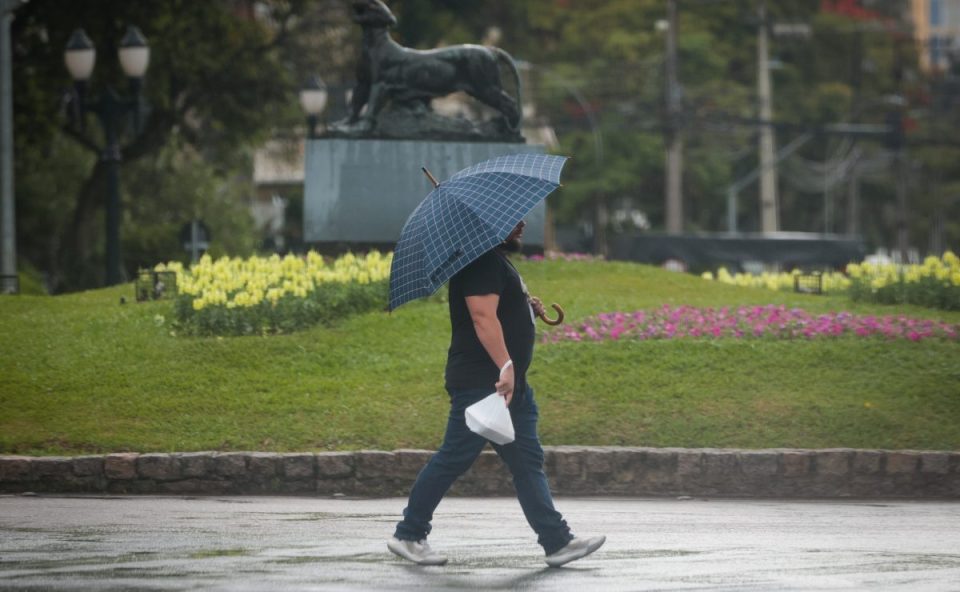 Imagem ilustra a previsão do tempo em Curitiba nesta terça-feira (30). Um homem de camiseta preta e calça jeans está caminhando na calçada, ao lado de um gramado verde, segurando um guarda-chuva aberto sobre a cabeça. O dia está chuvoso. Ele segura uma sacola branca na mão direita.