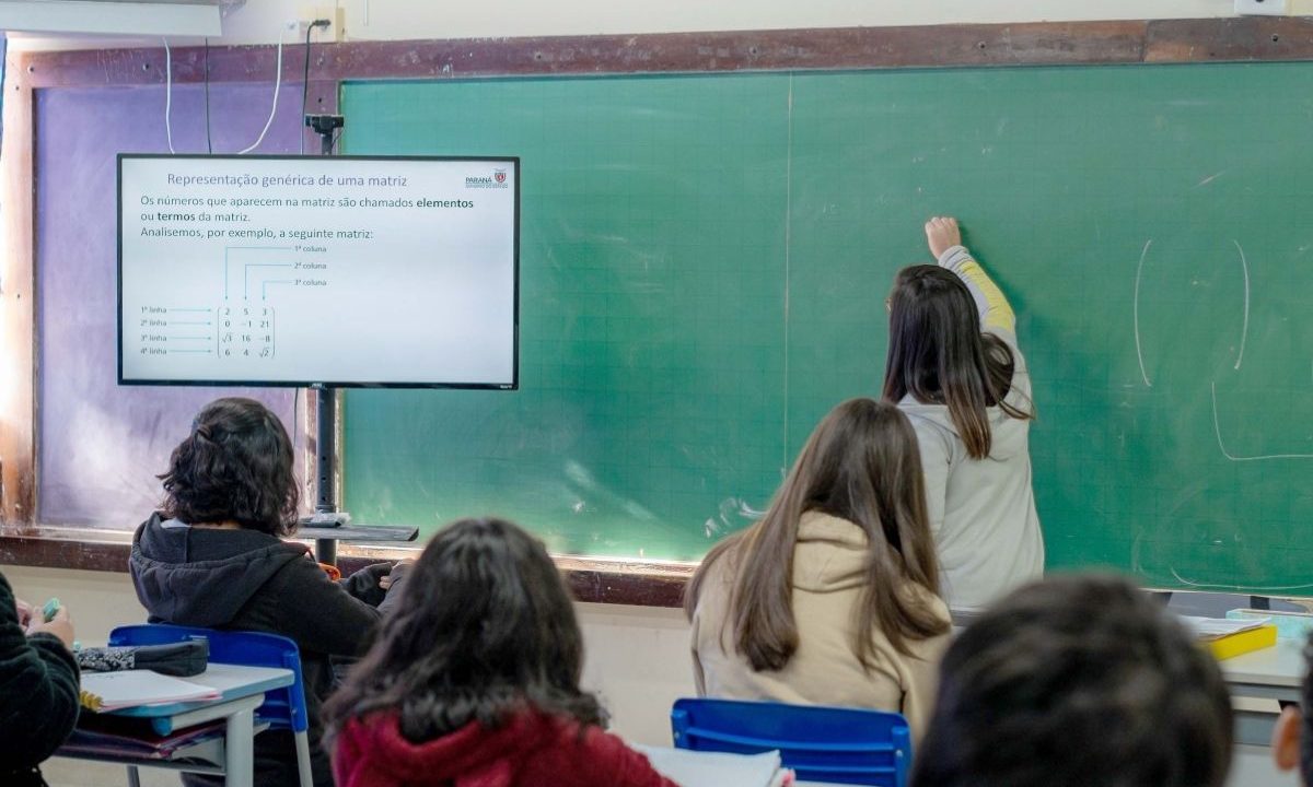 A imagem mostra alunos em sala de aula enquanto a professora escreve no quadro. A imagem representa professores da rede estadual do Paraná que, após avanço nas negociações com o Governo, adiaram o início da greve.