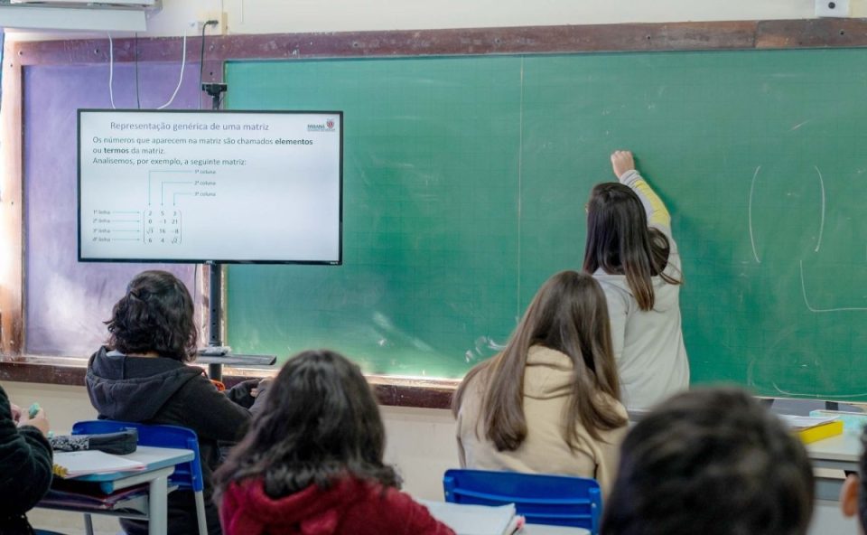 A imagem mostra alunos em sala de aula enquanto a professora escreve no quadro. A imagem representa professores da rede estadual do Paraná que, após avanço nas negociações com o Governo, adiaram o início da greve.