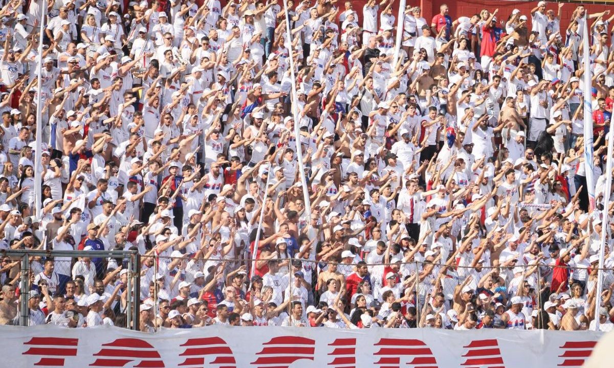 A imagem mostra uma arquibancada lotada de torcedores em um estádio de futebol, sob luz forte do sol. A maioria das pessoas veste camisetas brancas, criando um visual uniforme na multidão. Muitos torcedores estão com os braços levantados e estendidos para frente, como se estivessem cantando ou participando de uma coreografia coletiva da torcida. Alguns usam bonés, óculos escuros e seguram bandeiras ou faixas com cores vermelha e azul. É possível ver uma divisória de ferro com grades na parte inferior da imagem, separando setores do estádio. A cena transmite muita energia, com a torcida compacta, animada e engajada no apoio ao time.