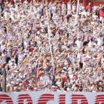 A imagem mostra uma arquibancada lotada de torcedores em um estádio de futebol, sob luz forte do sol. A maioria das pessoas veste camisetas brancas, criando um visual uniforme na multidão. Muitos torcedores estão com os braços levantados e estendidos para frente, como se estivessem cantando ou participando de uma coreografia coletiva da torcida. Alguns usam bonés, óculos escuros e seguram bandeiras ou faixas com cores vermelha e azul. É possível ver uma divisória de ferro com grades na parte inferior da imagem, separando setores do estádio. A cena transmite muita energia, com a torcida compacta, animada e engajada no apoio ao time.