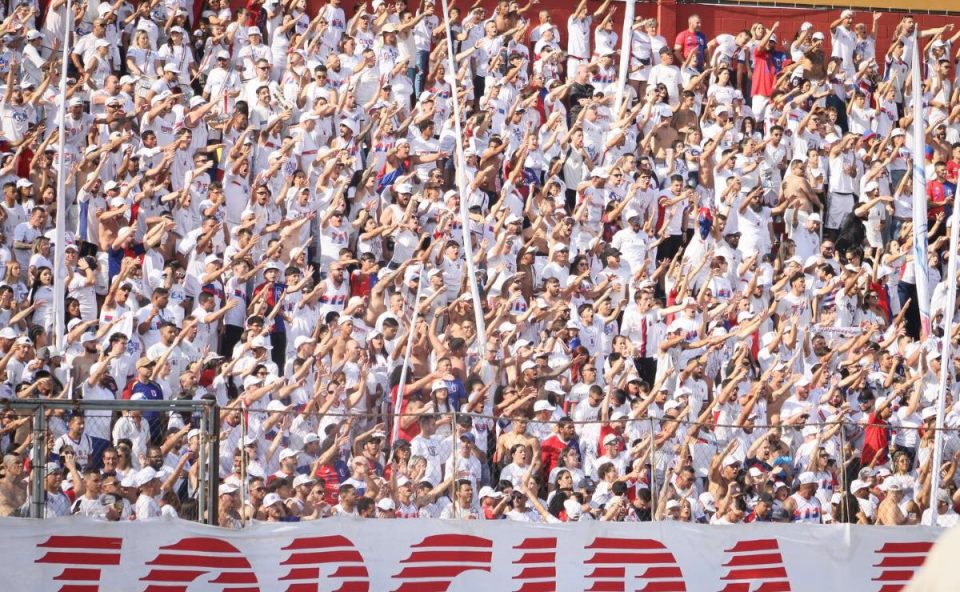 A imagem mostra uma arquibancada lotada de torcedores em um estádio de futebol, sob luz forte do sol. A maioria das pessoas veste camisetas brancas, criando um visual uniforme na multidão. Muitos torcedores estão com os braços levantados e estendidos para frente, como se estivessem cantando ou participando de uma coreografia coletiva da torcida. Alguns usam bonés, óculos escuros e seguram bandeiras ou faixas com cores vermelha e azul. É possível ver uma divisória de ferro com grades na parte inferior da imagem, separando setores do estádio. A cena transmite muita energia, com a torcida compacta, animada e engajada no apoio ao time.