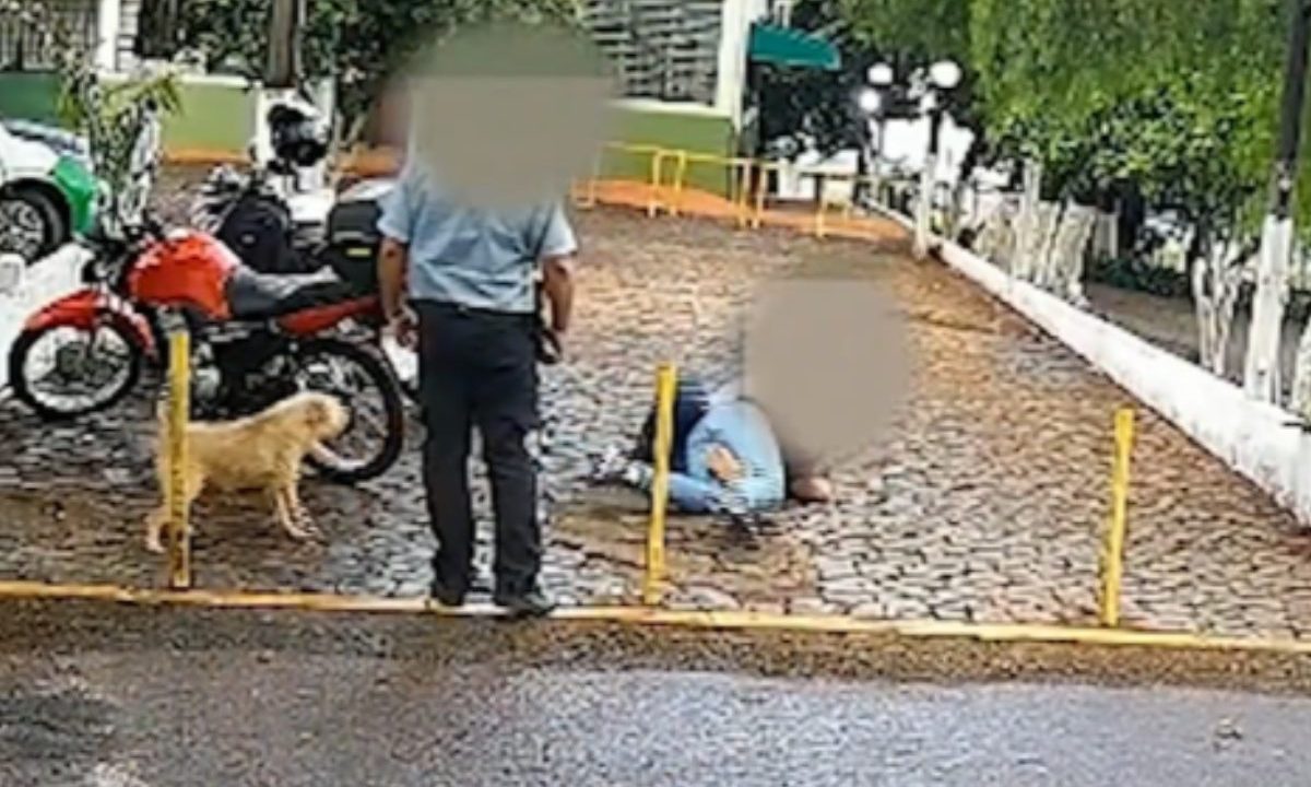 Segurança armado assiste assalto dentro de universidade. Imagem mostra o segurança parado olhando o crime. Dois homens estão no chão, perto de motos e de um cachorro caramelo.