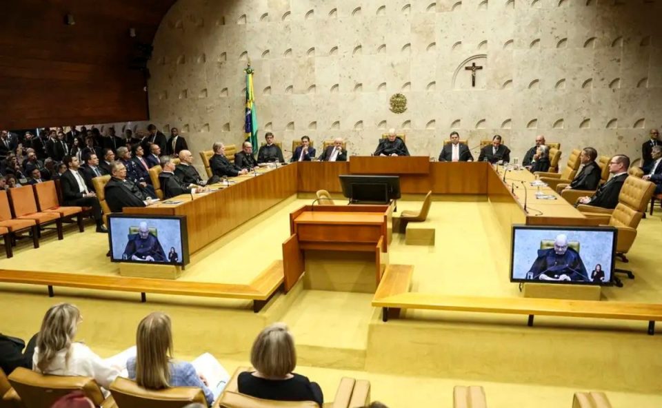 A imagem mostra ministros do STF sentados em suas cadeiras. Todos usando togas pretas. Ao fundo, uma parde em tons claros, assim como o chão.