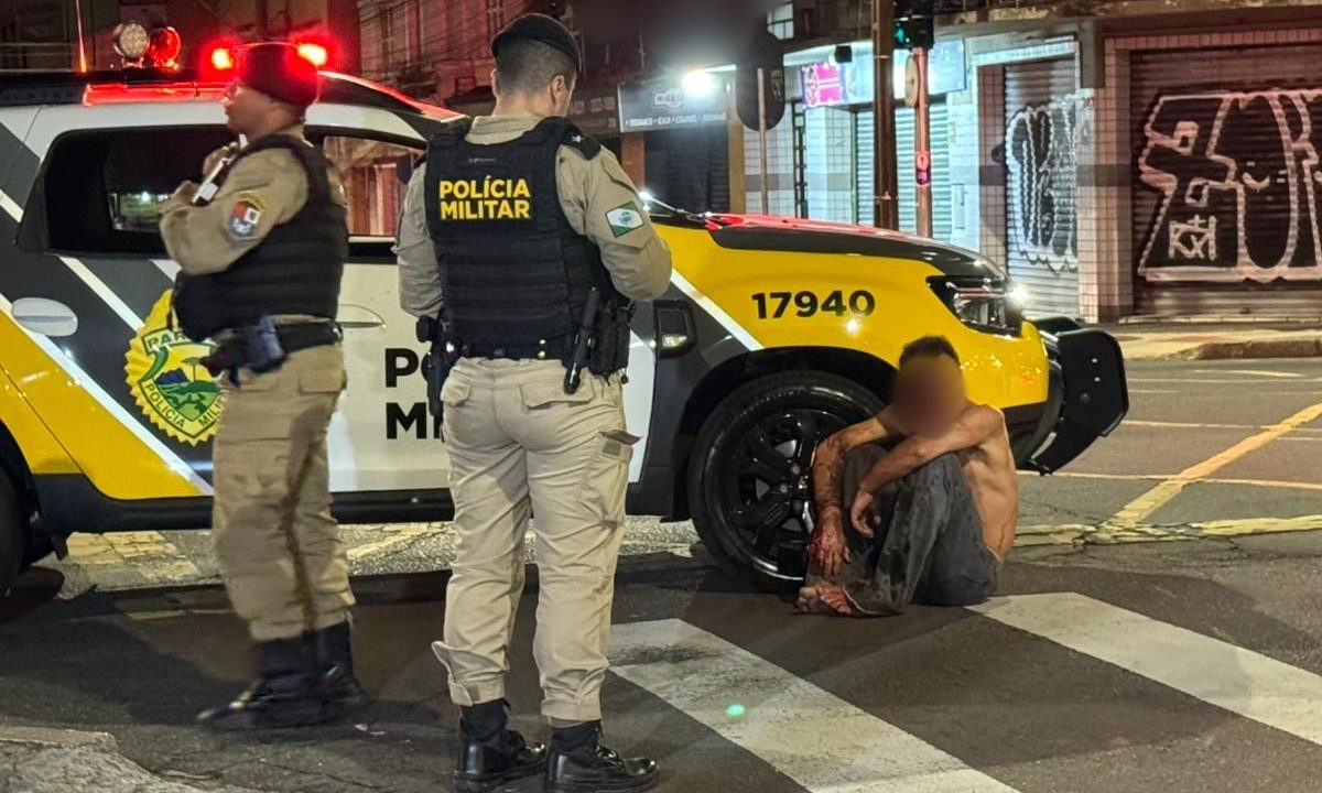 Homem espancado após suspeita de furto de celular aparece sentado no chão ao lado de viatura da Polícia Militar no Centro de Curitiba, durante atendimento da ocorrência na noite de terça-feira. Dois policiais militares fardados estão em frente à vítima, que está com o rosto borrado