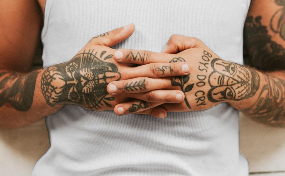 Imagem mostra um jovem com as mãos cruzadas. O braço e as mãos são tatuados. O tema é doença ocular que pode ser provocada por tatuagem. O jovem usa regata branca e aparece deitado.