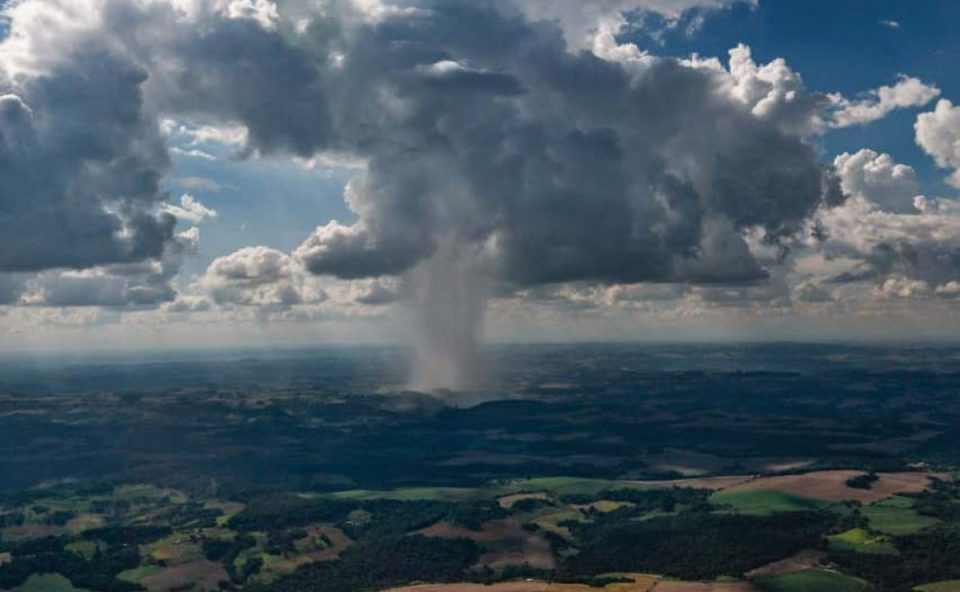 Imagem mostra o céu carregado de nuvens com tempo de chuva no Paraná. Embaixo aparece uma cidade em meio a área de mata e vegetação.