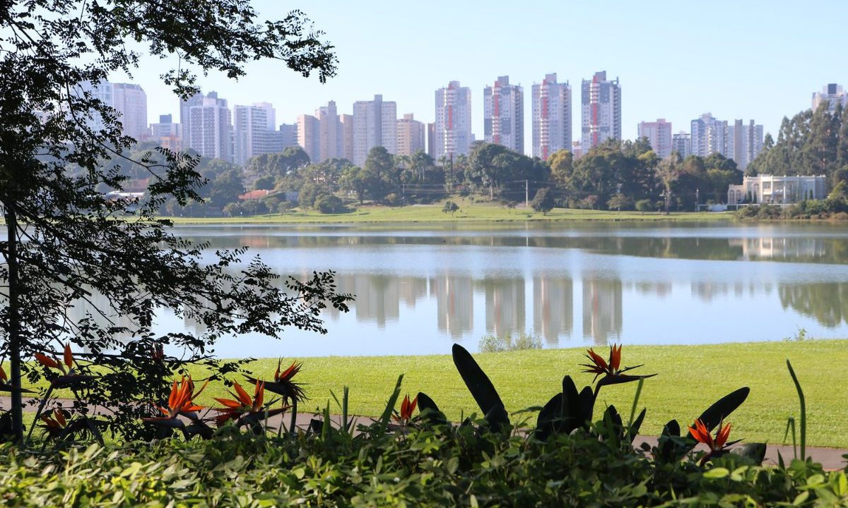 Imagem mostra o Parque Barigui em Curitiba, em um dia de sol e calor. A previsão do tempo para o fim de semana indica estabilidade. Imagem mostra um lago, perto de um gramado verde. Há folhas de árvores e flores perto da foto. Atrás, há prédios na cidade.