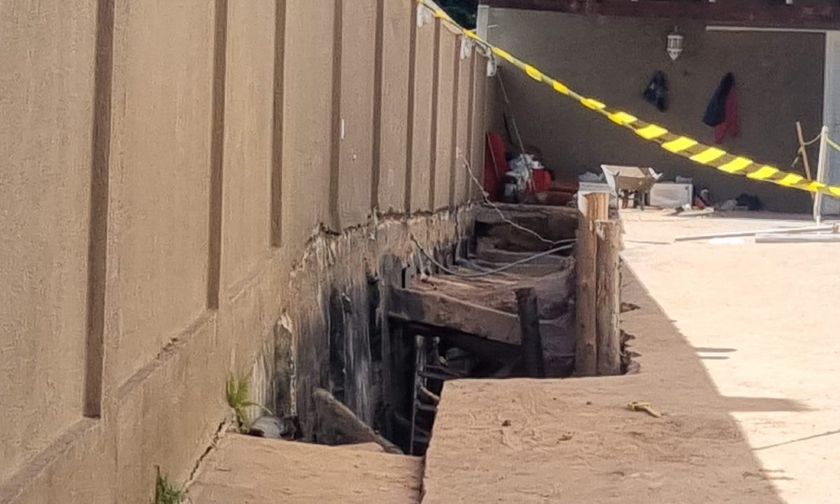 Área externa com muro danificado e parte da estrutura cedida, formando um buraco. O local está isolado com fita de segurança após desabamento no bairro Vista Alegre, onde trabalhador morreu soterrado.