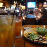 Mesa de bar com dois drinques gelados e prato de tacos com guacamole e queijo, com clientes desfocados ao fundo
