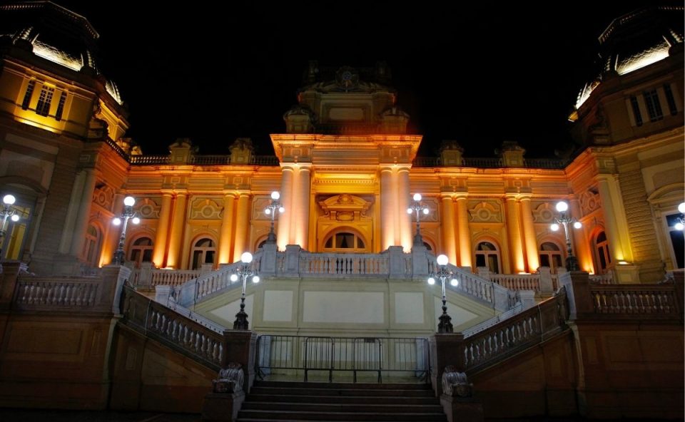 Fachada do Palácio Guanabara iluminada à noite em tons de laranja. A imagem mostra a escadaria principal e a arquitetura eclética da sede do governo do Rio de Janeiro.