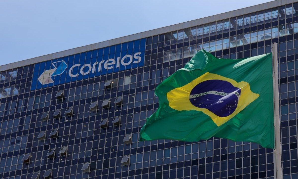Fachada do edifício-sede dos Correios em Brasília, com destaque para o logotipo azul e a bandeira do Brasil tremulando em primeiro plano sob um céu azul